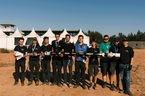 Ein Gruppenbild im Freien, die Gruppe hält gemeinsam eine Rakete, im Hintergrund weiße Zelte, der Boden wirkt sandig. 
