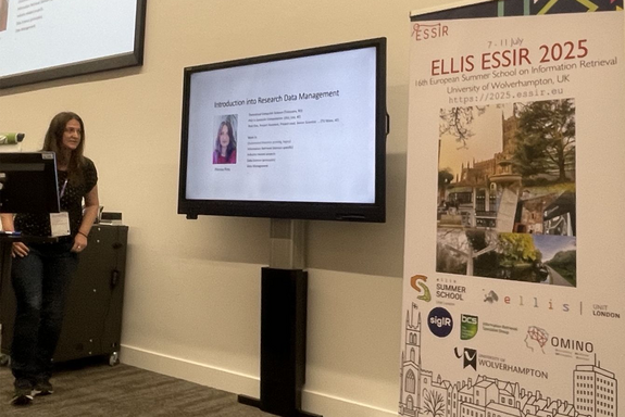 The photo shows a woman facing a presentation screen. On the right-hand side of the picture, a roll-up banner with information about the event can be seen.