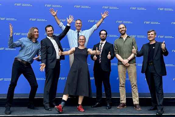 Sieben Personen posieren auf Bühne mit blauem Hintergrund.
