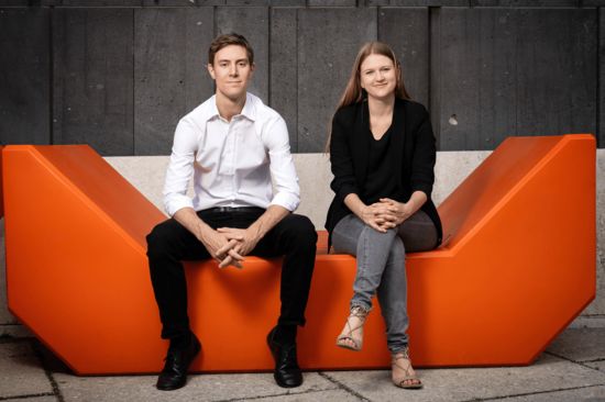 Die zwei Gründer_innen auf einem futuristischen orangen Sitzmöbel mit dunklem Hintergrund.