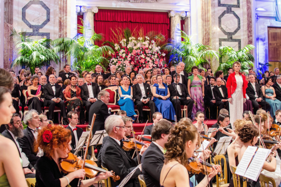 Festlich gekleidetes Orchester im Vordergrund, im Hintergrund Ballpublikum. Feierlicher Rahmen mit Palmendekoration.