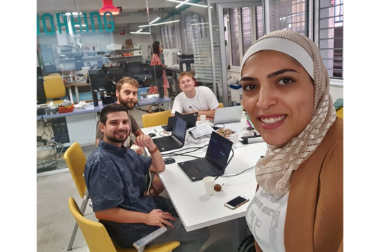 Das Bild zeigt Samah Jaber, die ein Selfie von sich und drei Kollegen macht, die mit ihren Laptops neben ihr an einem Tisch sitzen und in die Kamera schauen.