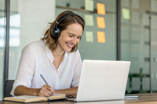 Eine lächelnde junge Frau mit Kopfhörern sitzt vor einem aufgeklappten Laptop und schreibt in ein Notizbuch.