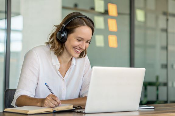A smiling young woman with headphones is sitting in front of an open laptop and writing in a notebook.