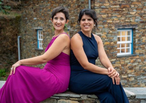 Elvire de Paiva e Pona (links), Joana Rolo (rechts) - beide Damen haben ein elegantes Abendkleid an, links lila, rechts dunkelblau. Sie sitzen mit dem Rücken zueinander.
