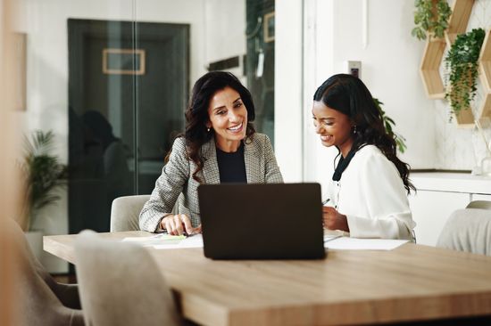 Two women are sitting together in front of a computer in an office and talking.