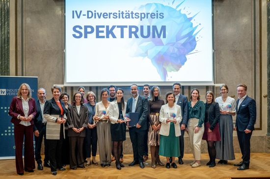 Gruppenfoto der Gewinner_innen des IV-Diversitätspreises Spektrum in einem festlichen Saal. Im Hintergrund sieht man eine Leinwand auf der "IV-Diversitätspreis SPEKTRUM" steht. 
