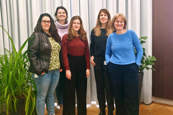 Gruppenbild mit fünf Personen vor einem Vorhang, die freundlich in die Kamera blicken, neutral gekleidet, alle tragen Hosen. Links daneben eine Pflanze.
