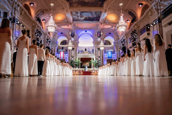 Balleröffnung durch die jungen Tänzer_innen. Frauen weiß gekleidet, Männer im schwarzen Anzug. Festsaal mit prächtigen Lustern und Deckenfresken.