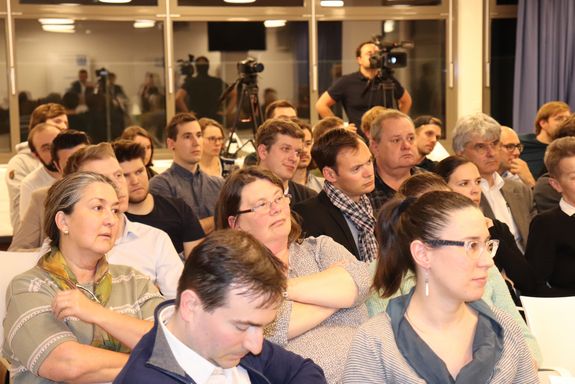 Ein Foto des Publikums. Auf dem Foto sind rund 30 Leute zu sehen. Im Hintergrund steht der Kameramann hinter seiner Kamera. Eine Fotokamera steht auf einem Stativ daneben. In den Fensterscheiben im Hintergrund spiegelt sich das Geschehen.