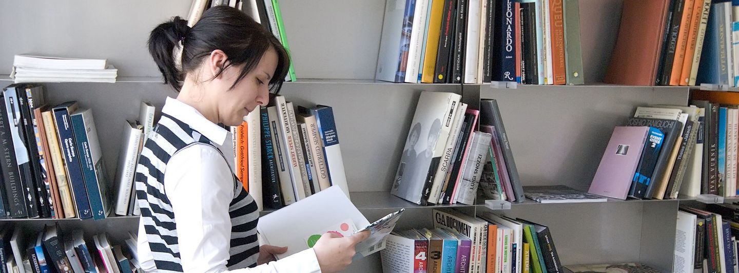 A woman with a book stands in front of a bookshelf