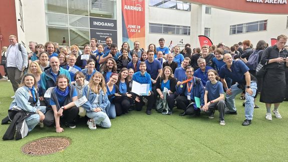 Erfolgreiche Sänger_innen nach der Verleihung der silbernen Medaille posieren vor einem Gebäude und strahlen in die Kamera. Sänger_innen sind blau-schwarz gekleidet.