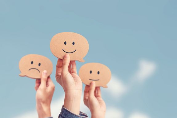 blauer Himmerl mit weißen Wolken im Hinetrgrund, davor drei Arme, die braune hölzerne Smileys halten