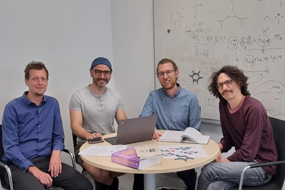 Vier Personen rund um einen Tisch sitzend, im Hintergrund eine Tafel mit Formeln