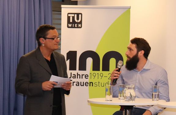 Christoph Kirchberger spricht in ein Mikrofon. Vor ihm ist ein hoher Tisch mit Wassergläsern und einem Wasserkrug. Links von ihm steht Norbert Fiala