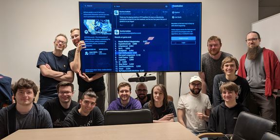 Gruppenbild des Teams KuK Hofhackerei nach der erfolgreichen Qualifikation für das Finale der DEFCON CTF. In der Bildmitte ist ein großer Bildschirm zu sehen. Darum und auf der Seite haben sich die Teammitglieder positioniert.