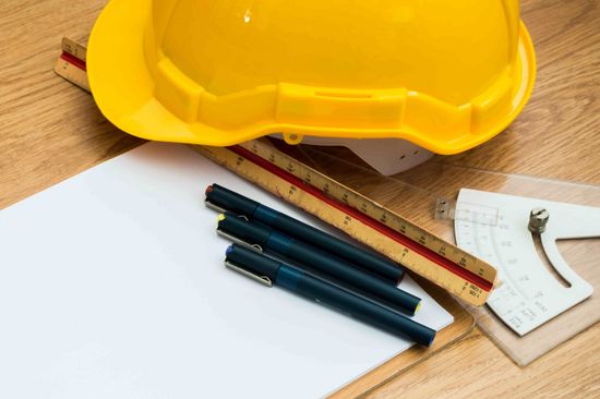 Auf Parkettboden liegen ein Klemmbrett, drei Stifte, zwei Lineale und ein gelber Schutzhelm