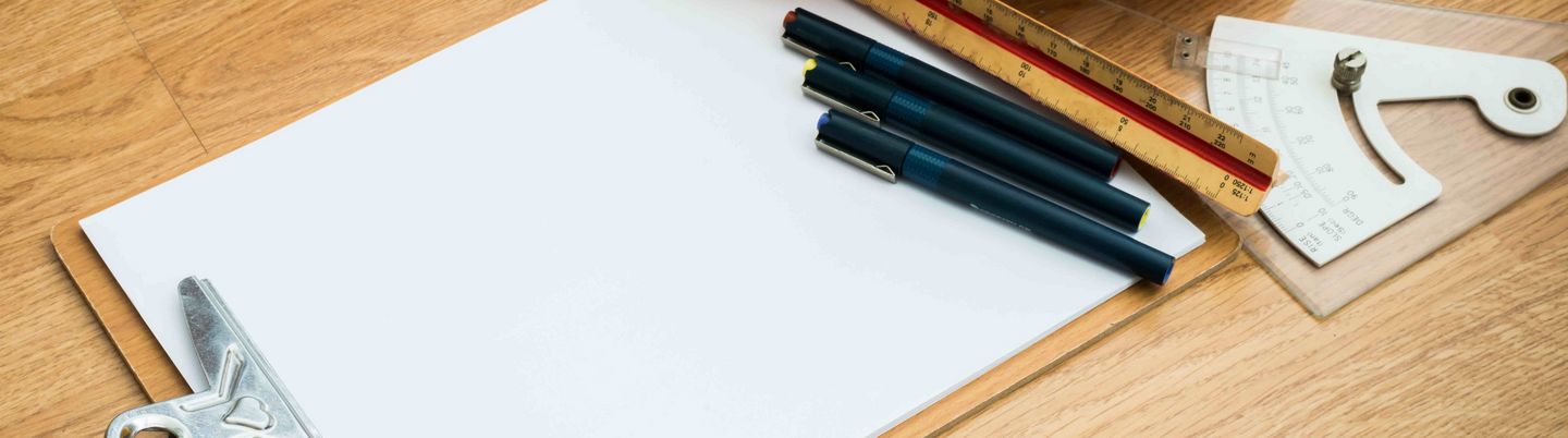 On a wooden floor there is a clipboard, some pencils, rulers and a helmet.