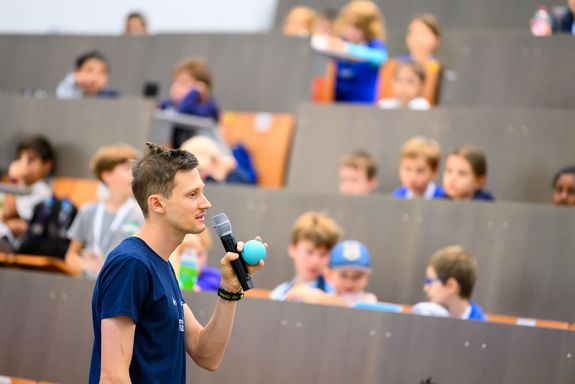 Kinder in Hörsaal, Vortragender Mann im blauen T-Shirt groß im Vordergrund