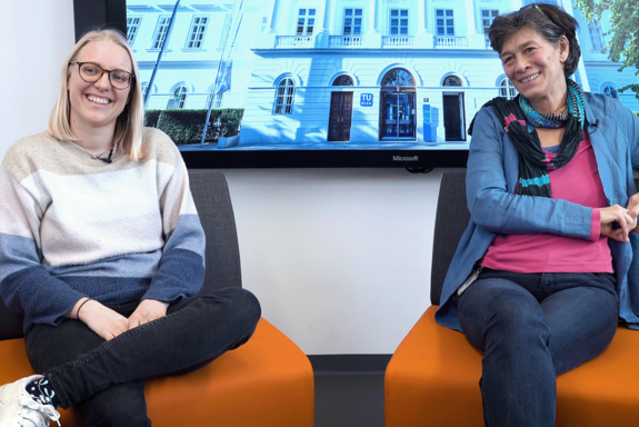 Dinah Gaffal und Brigitte Ratzer auf orangen Fauteuils sitzend. Im Hintergrund ein Screen mit dem Hauptgebäude der TUW.