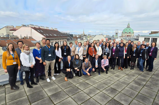 Gruppenfoto mit knapp 45 Personen auf einer Dachterrasse der TU Wien.  Im Hintergrund sind Dächer und die grüne Kuppel der Karlskirche zu sehen.