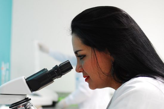 Junge, dunkelhaarige Frau in weißem Arbeitsgewand und mit Arbeitshandschuhen schaut ins Mikroskop. 