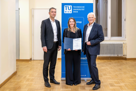 Drei Personen, in der Mitte die mit einer Urkunde ausgezeichnete. Alle in festlicher Kleidung mit einem TUW-Aufsteller im Hintergrund.