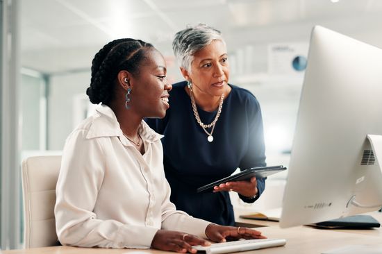 A photo of two women of different backgrounds in an office. One woman is explaining something to the other woman on the computer.