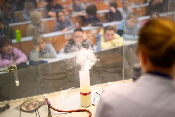 Frau in Labourmantel von hinten, vor ihr Versuchsaufbau mit rauchendem Glas