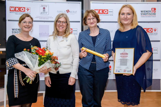 Gruppenfoto mit mit vier festlich gekleideten Personen, die jeweils einen Blumenstrauß, eine goldfarbene Rolle und eine Urkunde in Händen halten. Im Hintergrund Aufsteller mit "WKO" und "Wiener Städtische".