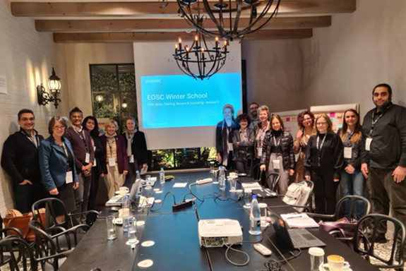 15 Personen stehen rund um einen Besprechungstisch vor einer Präsentationsleinwand.