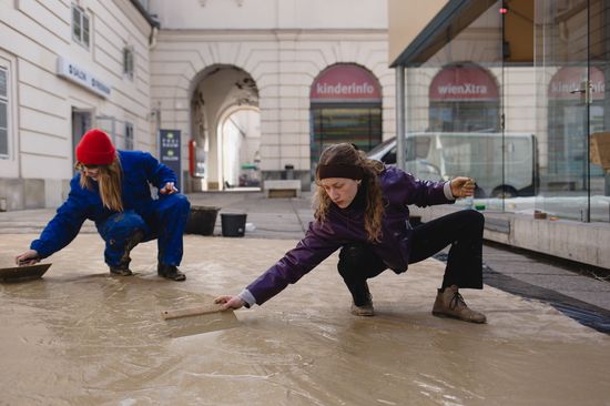 Zwei Studentinnen glätten einen Lehmboden im Hof der TU Wien