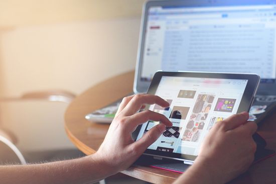 Webnews zwei Hände bedienen ein iPad auf einem runden Tisch. Im Hintergrund ist ein Computermonitor zu sehen.