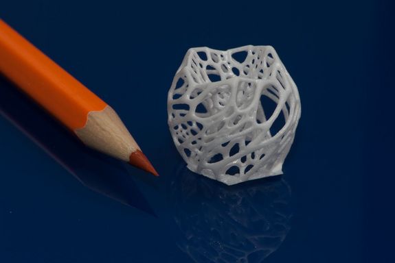 weißes, geometrisches Spritzgussteilchen, 2-3 Mal so hoch, wie der Buntstift der daneben liegt.