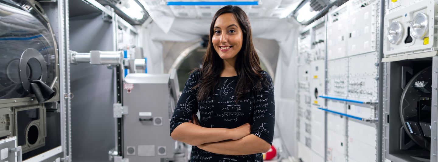 Selbstbewusste, junge Frau steht lächelnd in einem mit unterschiedlichen Maschinen ausgestatteten Raum. Sie trägt ein schwarzes Kleid mit vielen weißen, mathematischen Formeln. 