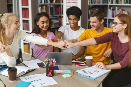 Five young people sit at a table full of study utensils and do a fist bump