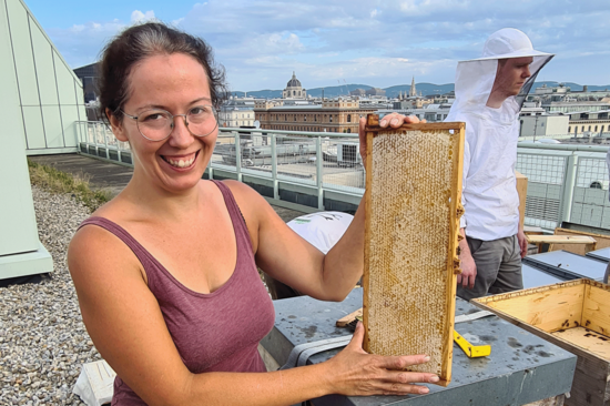 Imkerin Adriana neben einem Bienenstock, hält lächelnd eine voll gefüllte Wabe in die Kamera. Sie trägt ein lila Top. Im Hintergrund ein Impker mit Schutzkleidung, Ausblick auf die Dächer Wiens.