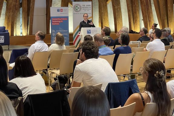 Vortrag, ein Sprecher auf Pult in der rechten Bildhälfte, davor Publikum sitzend