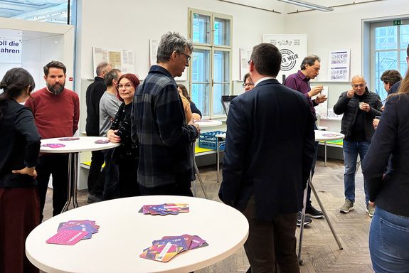 Im Vordergrund ein Stehtisch, auf dem punkte Pappuntersetzer liegen. Dahinter Menschen, die in Gruppen zusammen stehen und sich unterhalten.