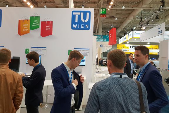 On the left in the "NGSolve" area and on the right in the "Epoxy Resin" area, interested people are standing talking.