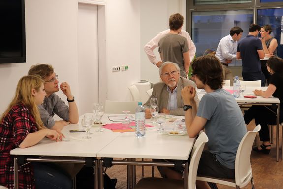 View of the audience. In the foreground four people are sitting at a table. In the background two more groups are lively discussing.