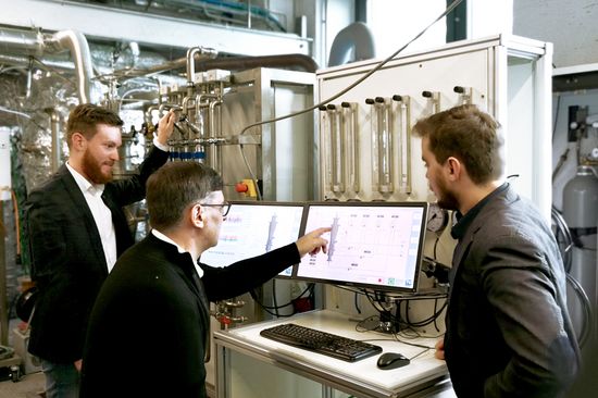 drei Personen vor einem Computerbildschirm und einer technischen Anlage mit zahlreichen Röhren