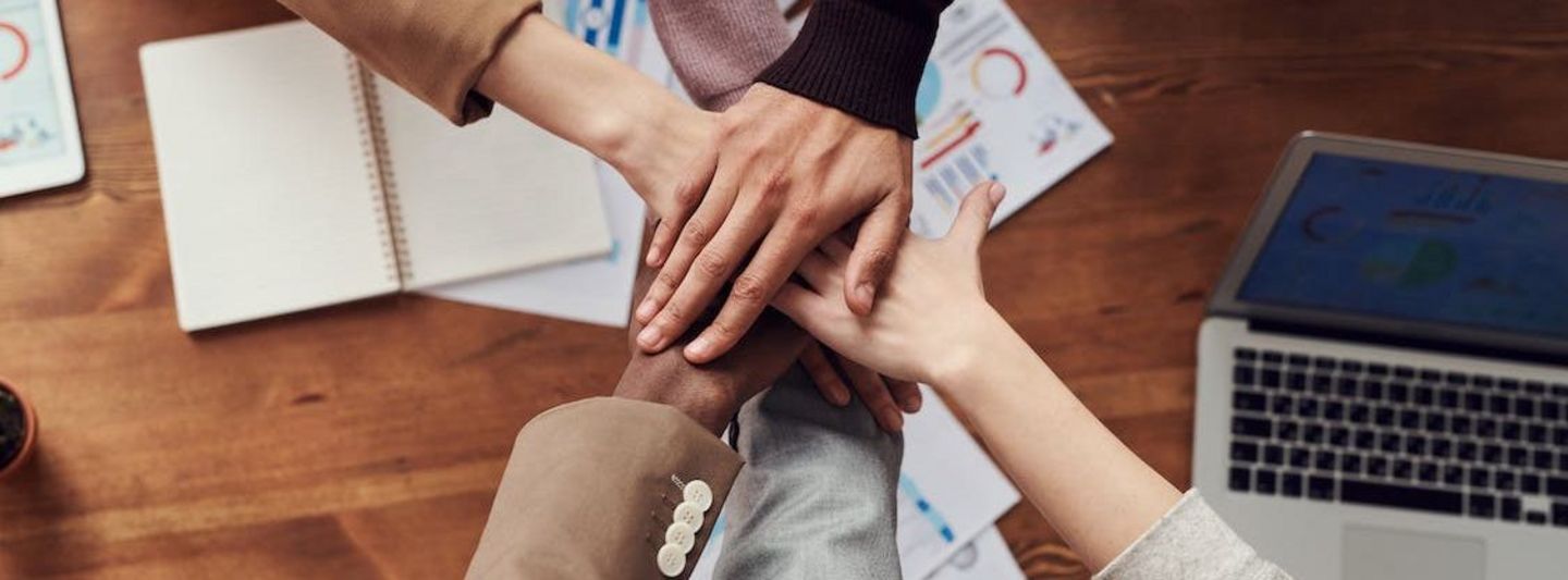 A top-down view of five people's hands coming together in a gesture of teamwork and unity over a wooden table, with visible papers containing graphs and a laptop, suggesting a collaborative work environment.