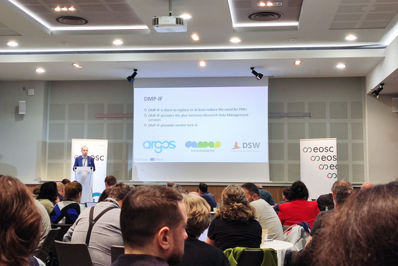 Das Foto zeigt einen Veranstaltungsraum mit Publikum, Vortragendem und Präsentationsleinwand.