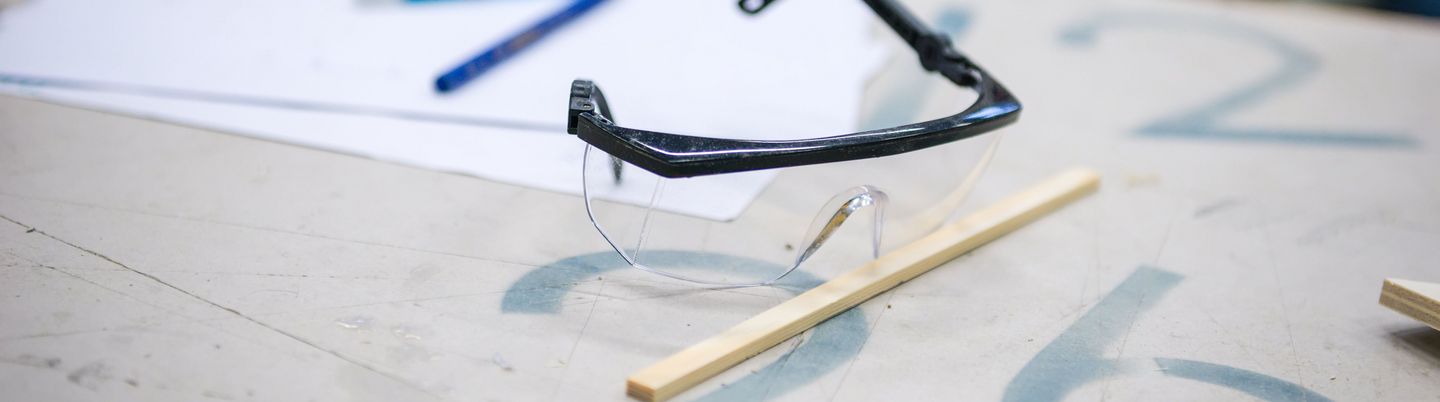Eine Schutzbrille liegt auf einem Tisch, davor ein Stück Holzverschnitt