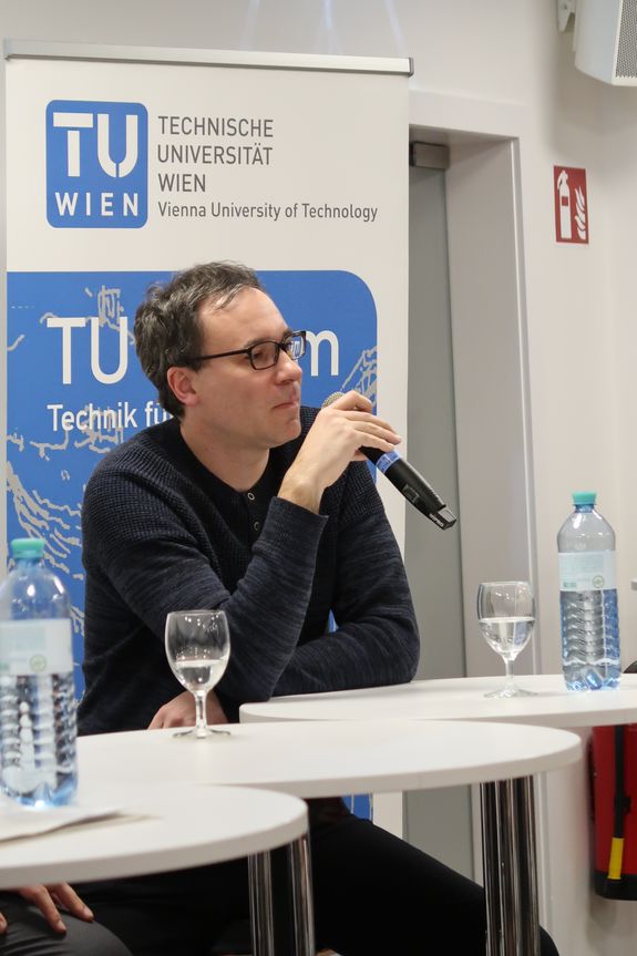Close-up by Thomas Müller. He sits at a high table and holds a microphone in his hand. In the background you can see a roll-up of TU Wien.