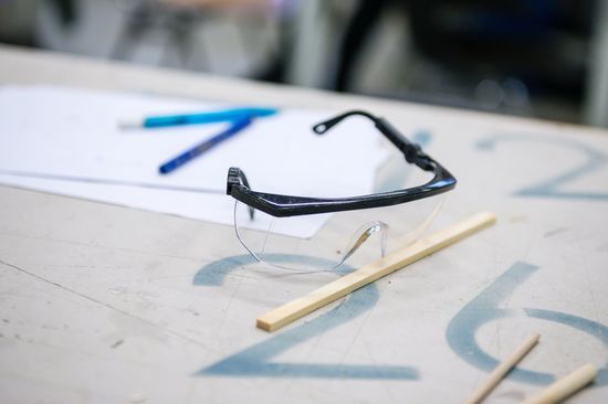 A pair of safety goggles lies on a table, behind it two blue pens and a sheet of paper