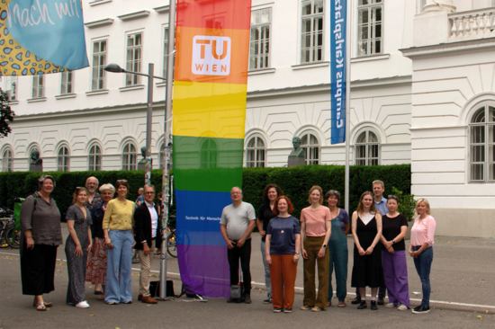 Gruppenbild vor dem TUW-Hauptgebäude mit einer Regenboggenflagge, auf der ein TUW-Logo abgebildet ist.