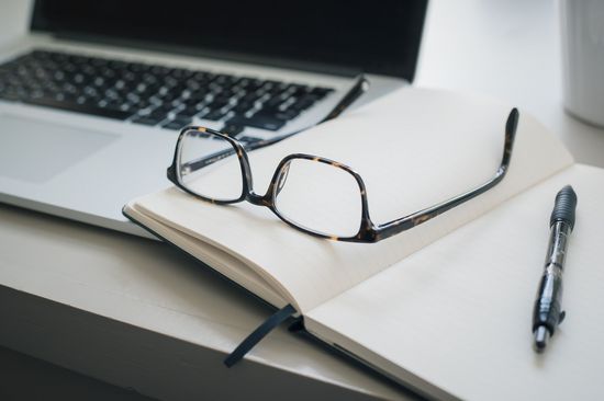 Laptop, Brille, Notizbuch und Stift liegen auf einem Tisch. 