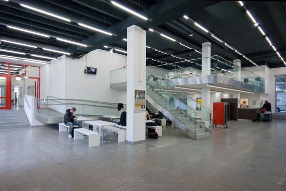 Aula - Mensa und Lernbereich Innenraum: große Geschoßhohe, rechts ist der Raum zweigeschossig. Die Treppe ist mittig platziert.